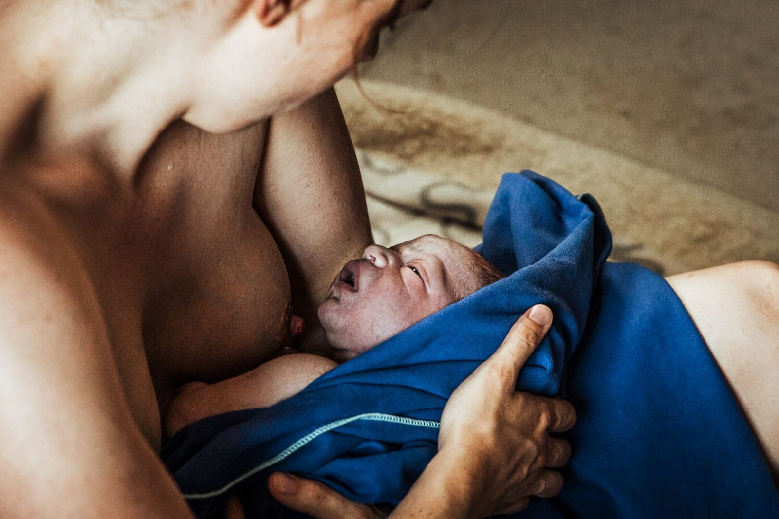 Wenige Minuten nach der Geburt. Mutter hält ihr Neugeborenes in einem blauen Tuch gewickelt an ihrer Brust und schaut es an.